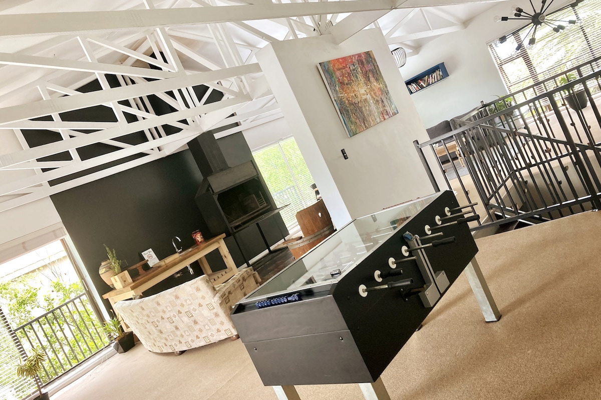 An open living area features a distinctive black wall and a modern foosball table, inviting friendly competition. A cozy seating area includes a comfortable sofa and a fireplace for warmth. Exposed beams and large windows ensure a bright and airy environment.
