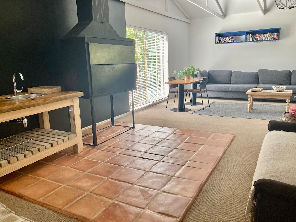 A well-lit indoor braai area features a sturdy black barbecue unit positioned against a dark wall. Natural light filters through large windows, illuminating a cozy lounge area with a beige carpet, a wooden dining table, and a grey sofa adorned with decorative cushions.