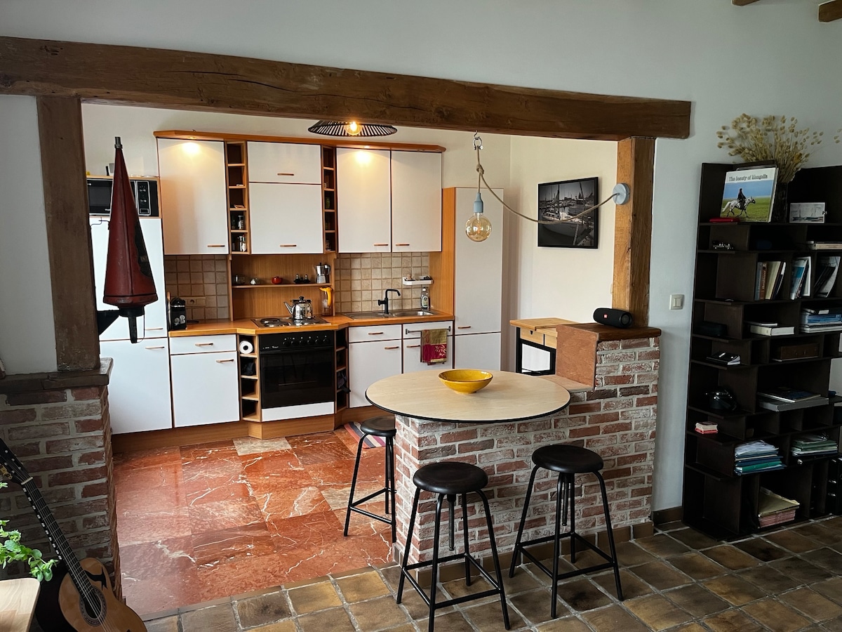 A modern kitchen features a spacious island with three black bar stools. The cabinetry is a combination of white and wooden finishes, while a brick accent wall adds warmth. Appliances are neatly arranged, with a coffee maker and a stylish light fixture illuminating the area.