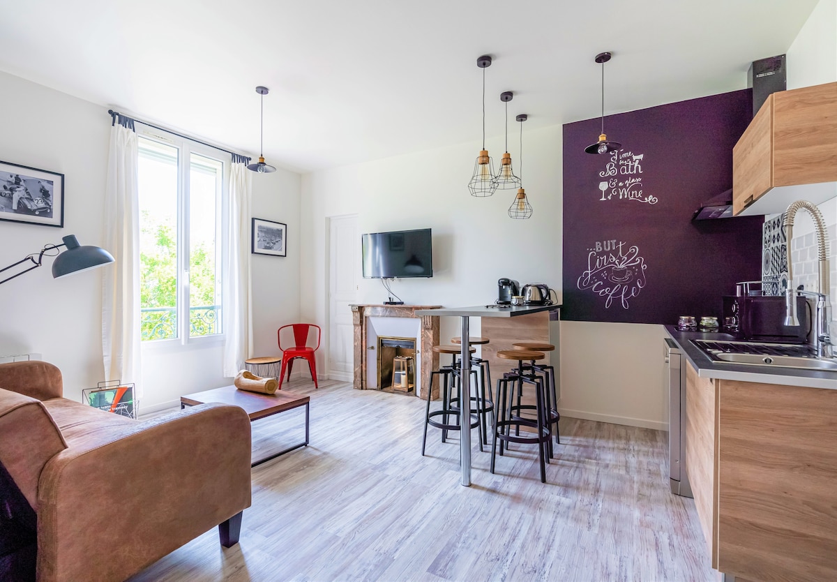 A modern living space with a comfortable seating area featuring a brown sofa and a small coffee table. A flat-screen TV is mounted on the wall opposite. A dining table with high stools is placed near the kitchenette, which includes kitchen appliances against a dark accent wall.
