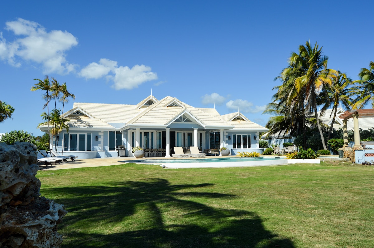 The oceanfront villa features a light blue exterior with a spacious lawn, bordered by palm trees. Large windows and a covered porch with seating are visible, providing natural light to the interior. A swimming pool is situated in front, highlighting the inviting outdoor space.