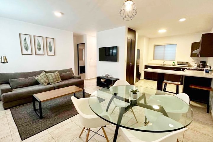 A modern living area is depicted, featuring a comfortable sectional sofa in neutral tones, a circular glass dining table with white chairs, and wall art with leaf designs. A flat-screen TV is mounted on the wall, while the kitchen area displays sleek cabinetry and appliances.