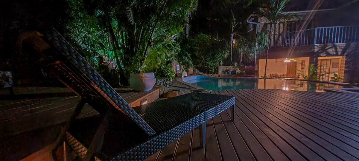 A serene night view of the designer swimming pool is framed by lush greenery, with soft lighting illuminating the surroundings. A comfortable lounge chair is positioned on the wooden deck, offering a spot to relax by the water.