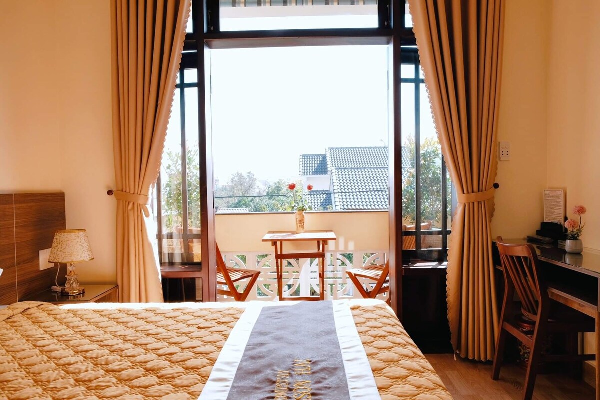A private room features a spacious bed adorned with a decorative blanket. Natural light streams through large windows, highlighting a wooden table and two chairs on the balcony. Soft curtains frame the space, contributing to a warm ambiance.
