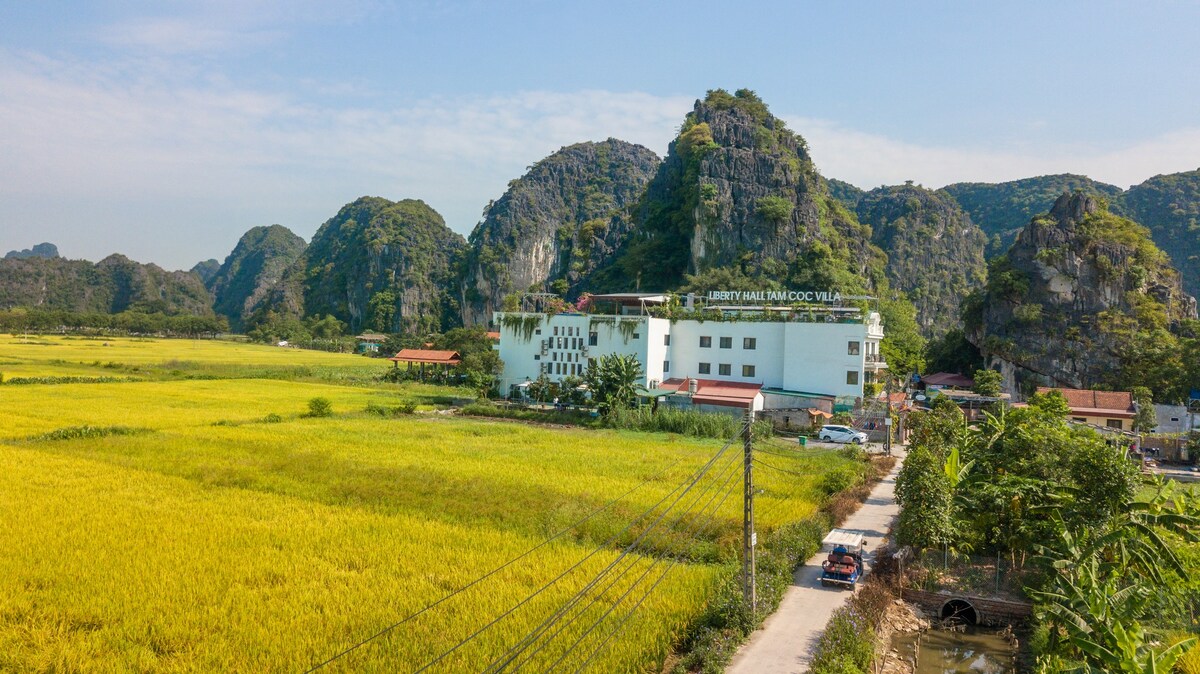 Aerial view of the hotel surrounded by vibrant green paddy fields and limestone mountains. The property is positioned along a narrow road, with a landscaped area in the foreground and a clear blue sky above, showcasing the natural beauty of the Ninh Binh region.