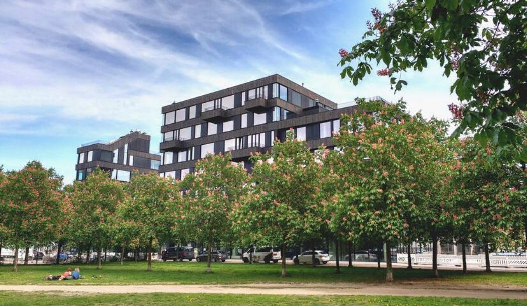 The exterior of the apartment building is showcased, set against a backdrop of a clear blue sky. The structure features a modern design with large glass windows. Lush trees with blooming flowers line the foreground, creating a green space that enhances the overall view.