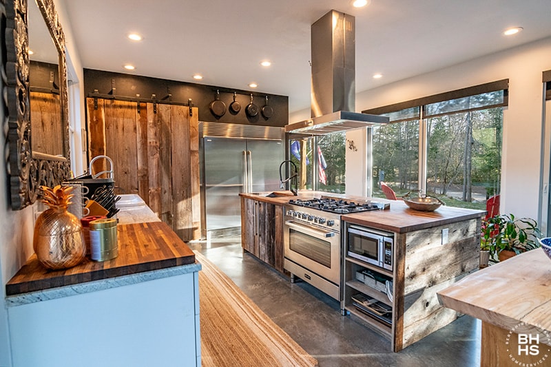 The kitchen features a central island with a wood countertop and stainless steel appliances, including an oven and vent hood. The walls showcase natural wood accents, and large windows allow for ample sunlight. Cooking utensils are displayed, adding functionality to the spacious design.