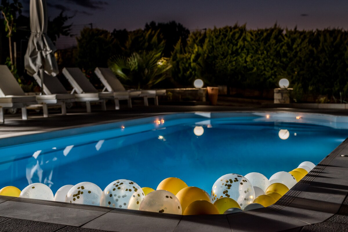 The illuminated private pool is surrounded by a stone deck, featuring white lounge chairs. Colorful floating balloons in white and yellow gently rest along the pool's edge, reflecting the evening lights and creating a cheerful ambiance.