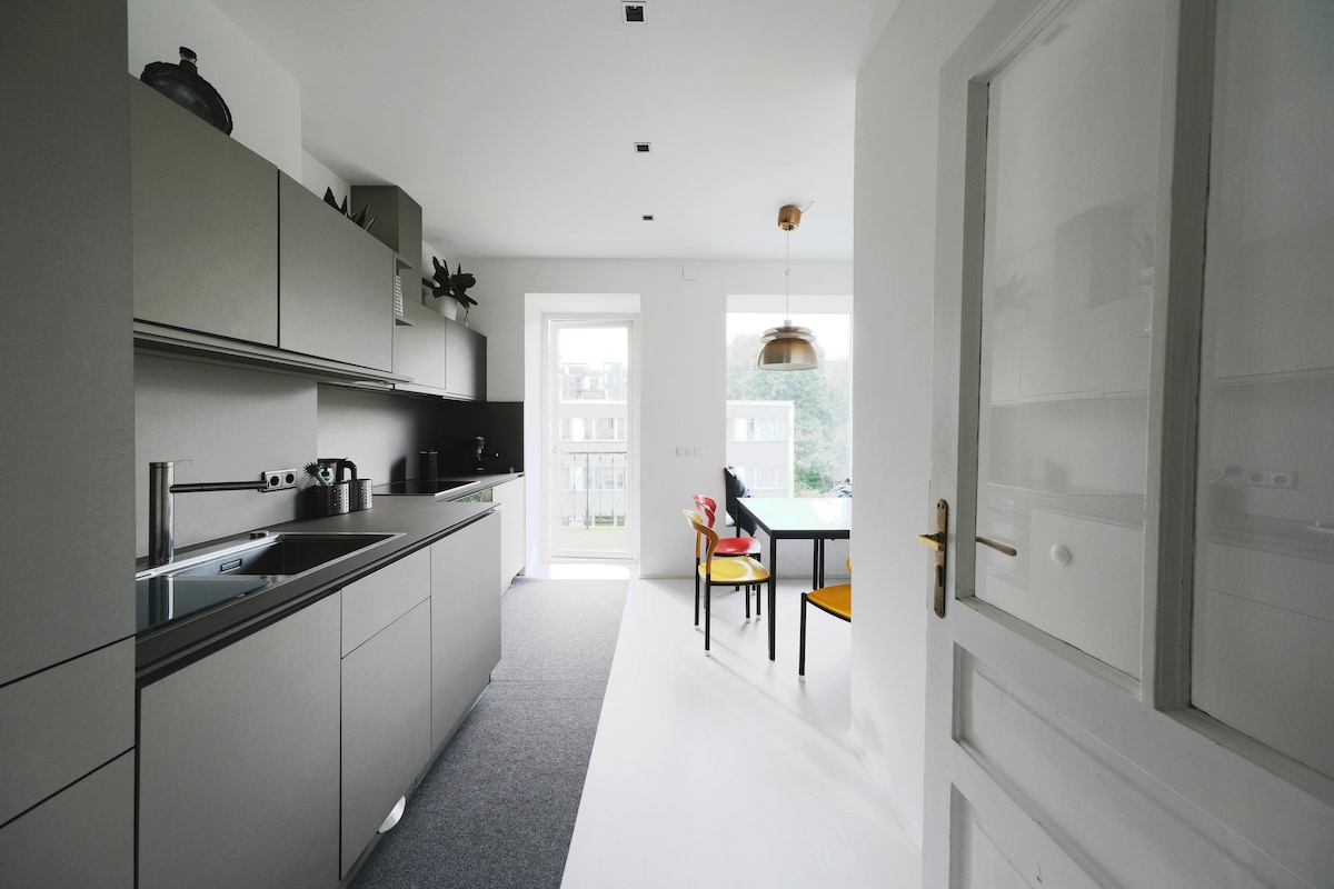 A modern kitchen showcases sleek cabinetry and a spacious counter. Natural light fills the area through a glass door leading to the balcony. A dining table with colorful chairs is positioned near the door. Various kitchen appliances and utensils are visible on the counter.