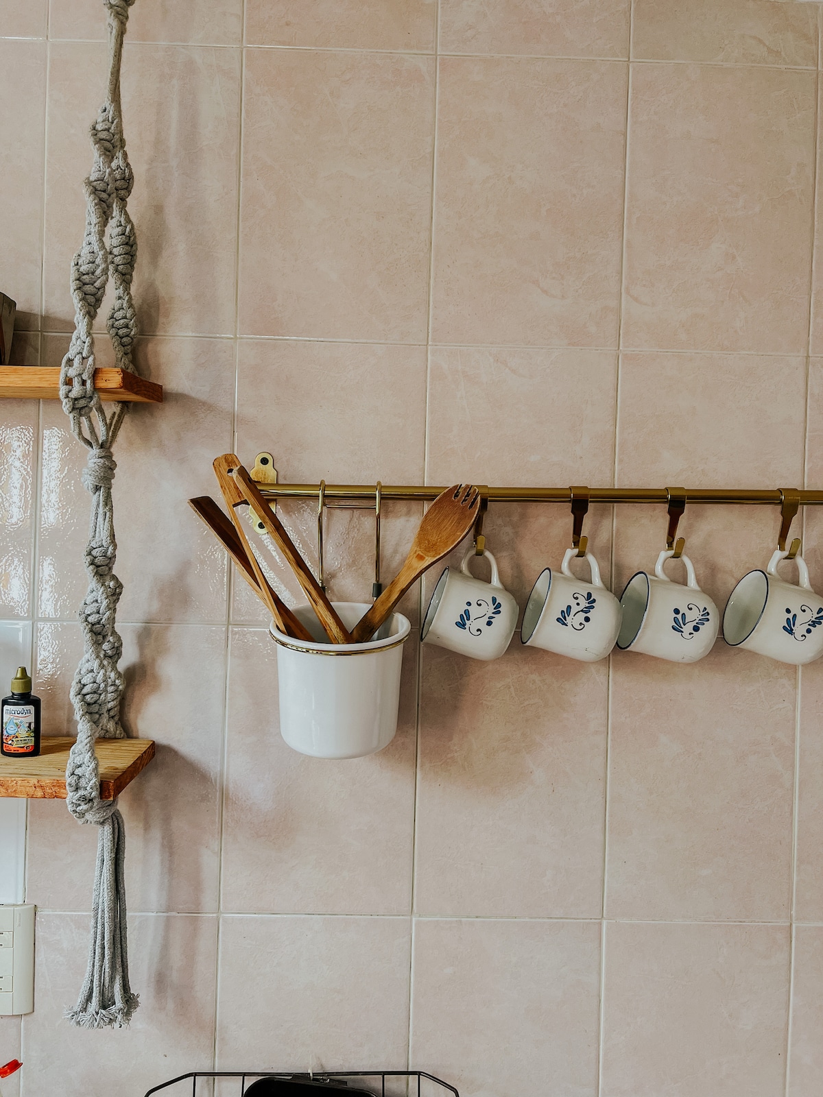 A well-organized kitchen area features hanging mugs adorned with a delicate design, alongside a container filled with wooden utensils. Light-colored tiles provide a bright backdrop, complemented by wooden shelves displaying macrame art and cooking essentials.