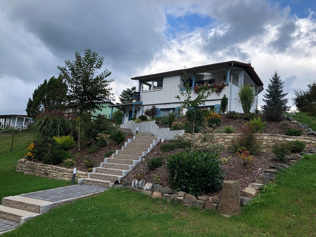 The house is situated on a hillside, surrounded by landscaped gardens featuring various plants and flowers. Stone steps lead up to the entrance, and windows are visible on the upper level, reflecting the natural light and scenery.
