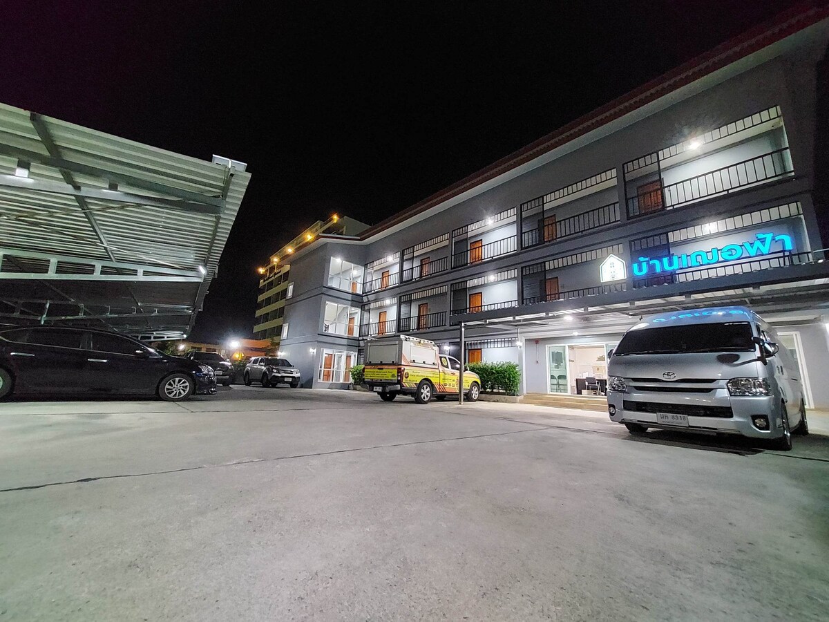 A night view of the property shows a multi-story building with lit balconies and a well-maintained exterior. The parking area accommodates several vehicles, including two parked vans and a small yellow vehicle. Soft lighting enhances the welcoming ambiance.