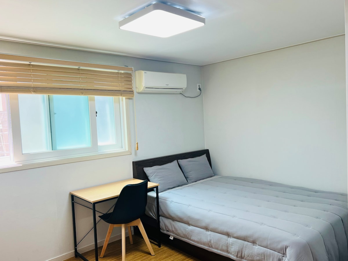 A bright and clean room features a spacious bed with gray bedding and a simple wooden desk paired with a black chair. Natural light enters through a window with blinds, while a ceiling air conditioning unit is mounted for comfort.
