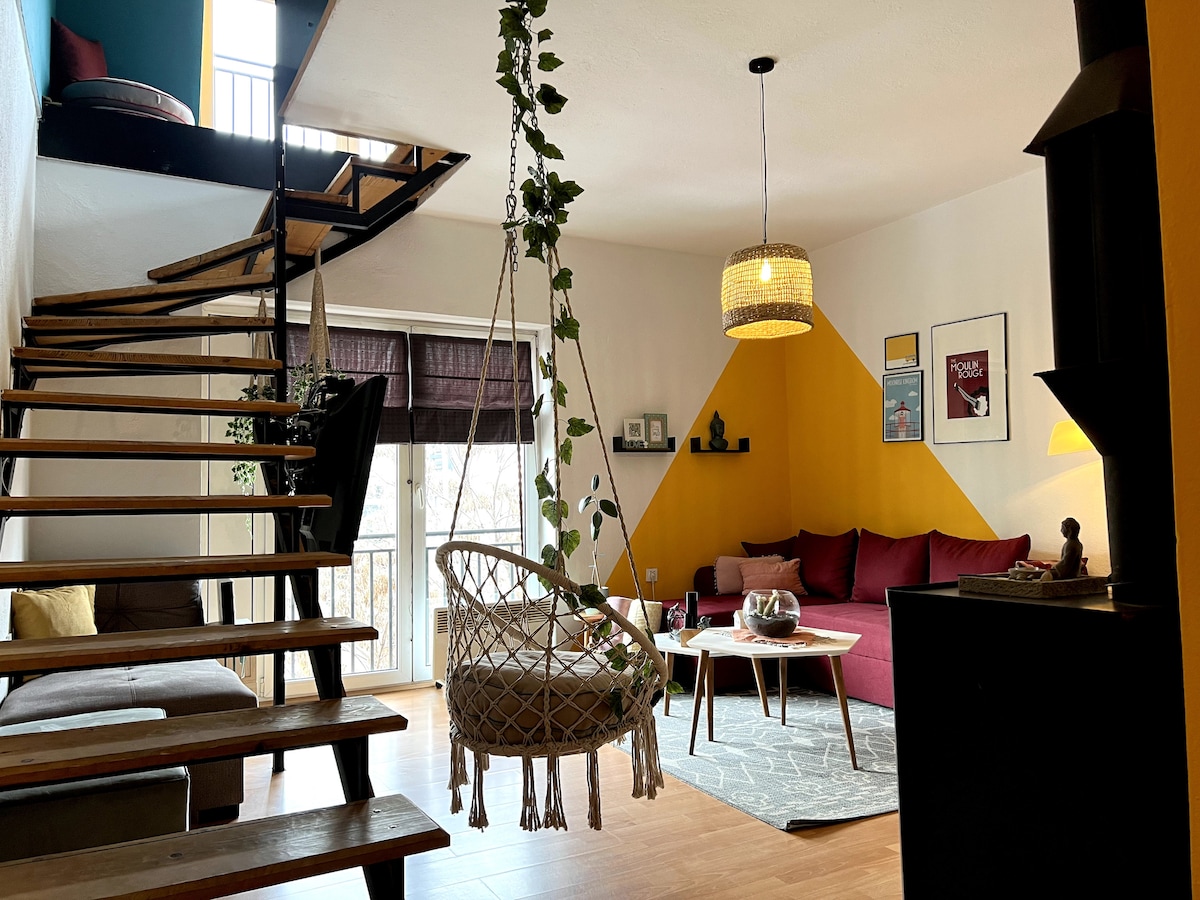 A cozy living room features a yellow accent wall, a comfortable seating area with a maroon sofa, and a glass coffee table positioned on a patterned rug. A hanging chair is suspended near a staircase leading to the upper level. Natural light enters through large windows with dark coverings.
