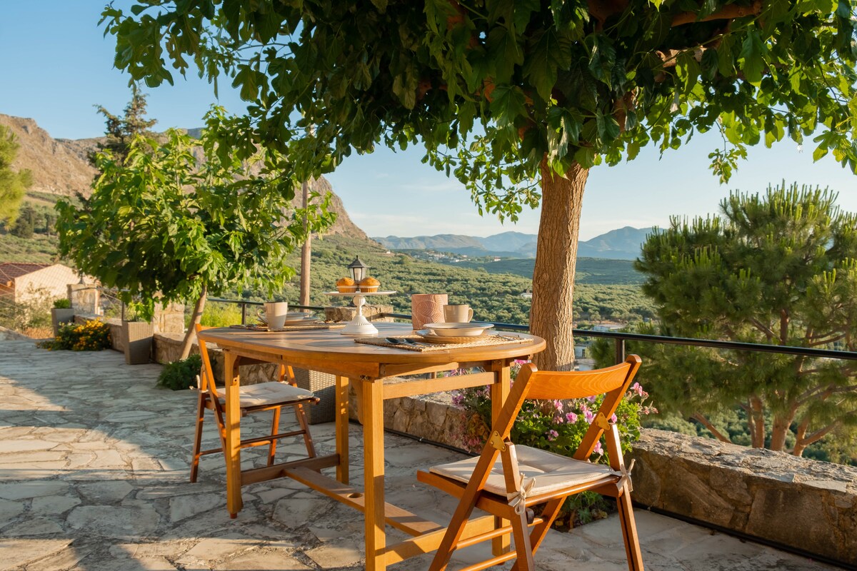 A wooden dining table is set under the shade of a leafy tree, surrounded by scenic mountain views. Two folding chairs are placed at the table, with a plate arrangement visible. Bright greenery and flowers enhance the tranquil outdoor environment.
