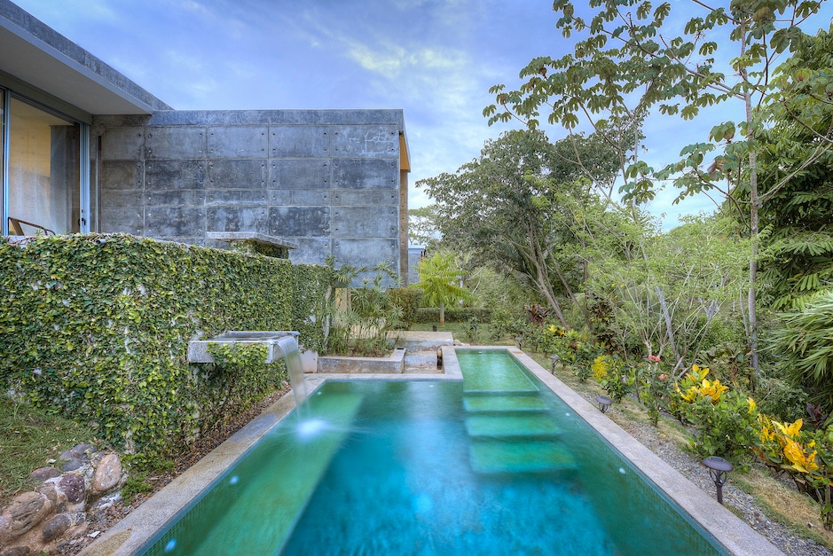 A private pool with varying depths is surrounded by lush greenery and tropical landscaping. The sleek concrete structure provides a modern backdrop, blending seamlessly with the natural environment. The water shimmers under the serene sky, inviting relaxation and tranquility.