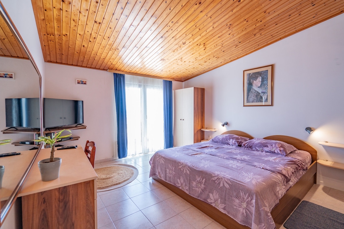 A spacious sleeping area is highlighted by a king size bed with a lavender bedspread. Natural light flows in through glass doors leading to a private balcony. A large mirror and a smart TV are visible on the opposite wall, complemented by a simple desk and chair.