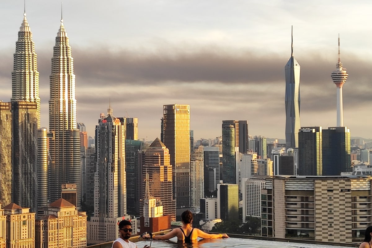 An expansive city skyline is showcased at sunset, featuring the iconic KLCC Petronas Twin Towers and the KL Tower. Two individuals are seen relaxing by an infinity pool, offering a stunning view of the cityscape, with warm light reflecting off the buildings.