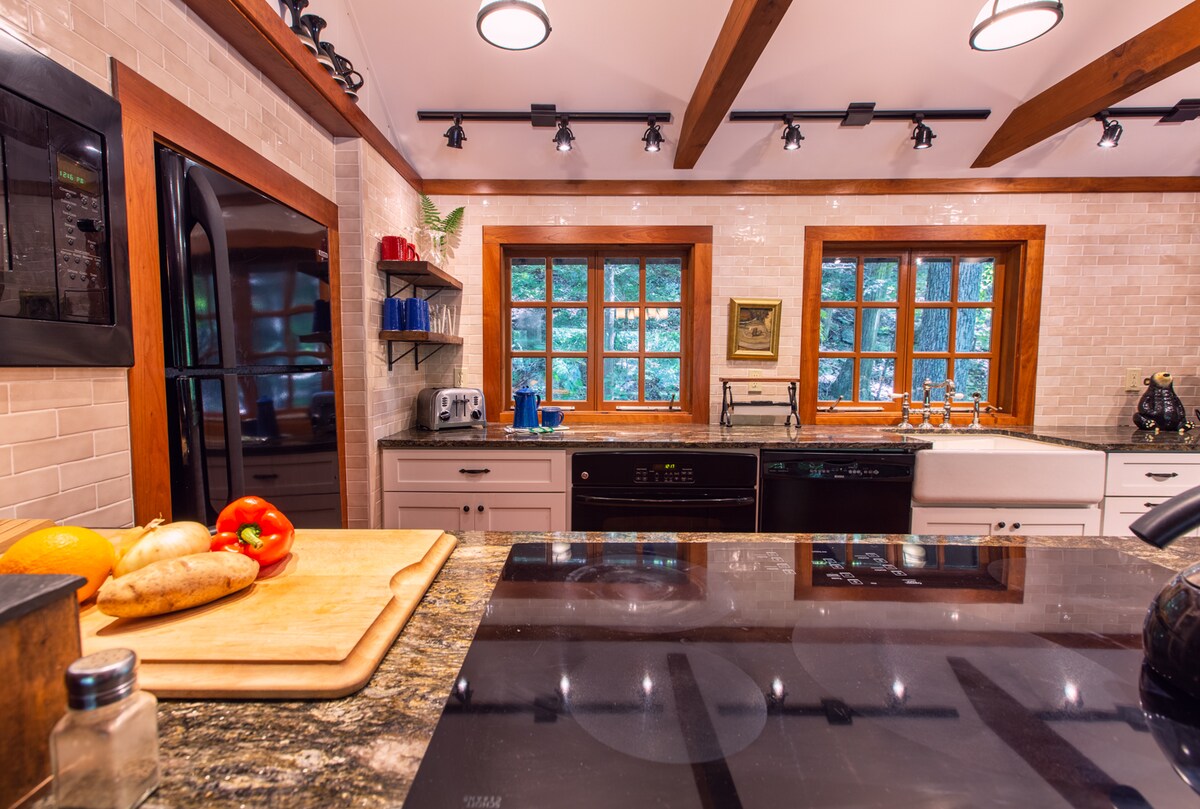 The modern kitchen features granite countertops with ample preparation space and a wood cutting board displaying fresh vegetables. Black appliances are positioned against white cabinetry. Four windows invite natural light, providing views of the surrounding greenery. Additional shelving holds kitchenware and a small appliance.