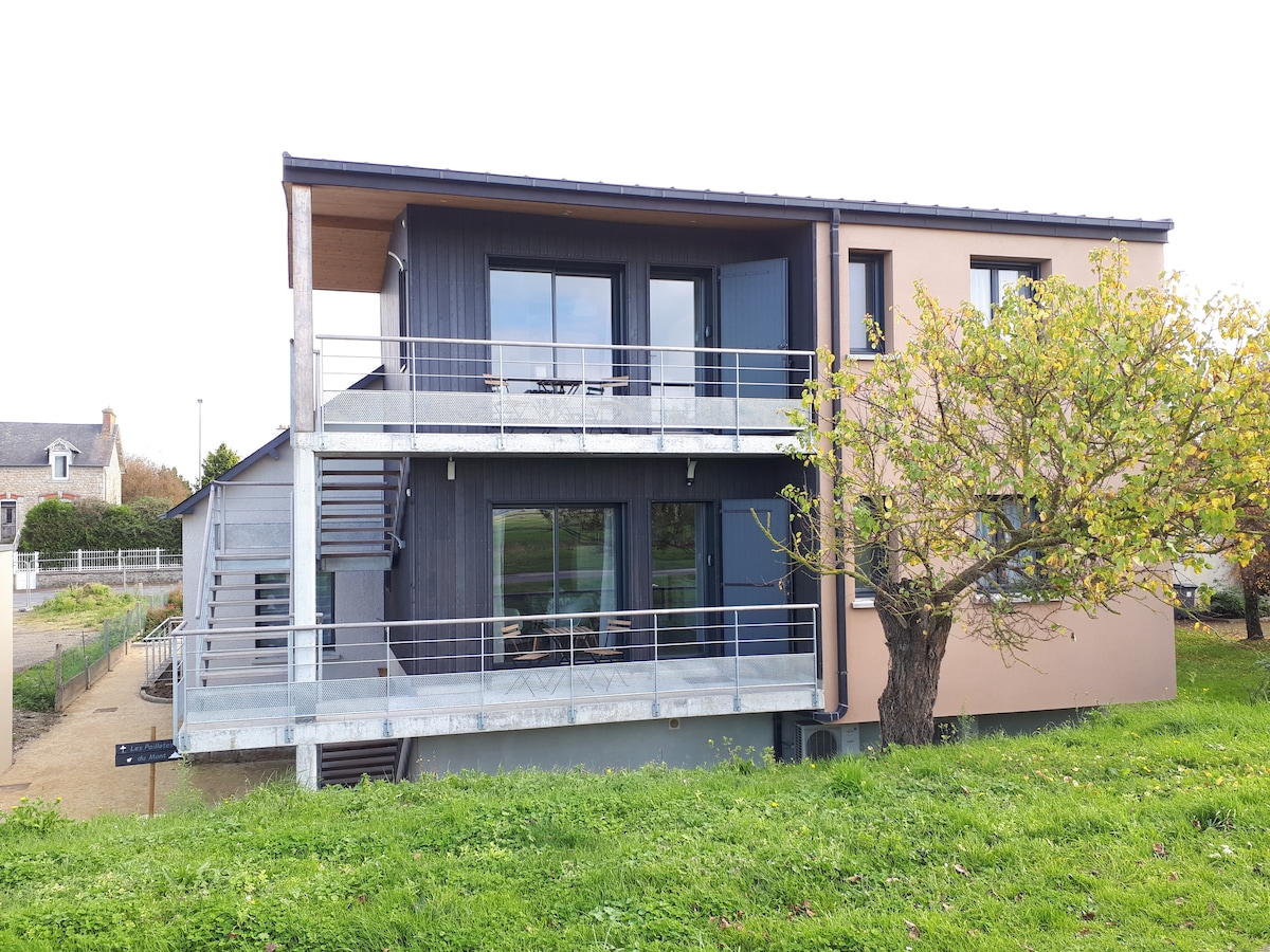 The exterior of the accommodation is shown, featuring a two-story modern structure with large windows. A balcony with railings extends from the upper level. A tree and grassy area are visible in the foreground, creating a serene setting.