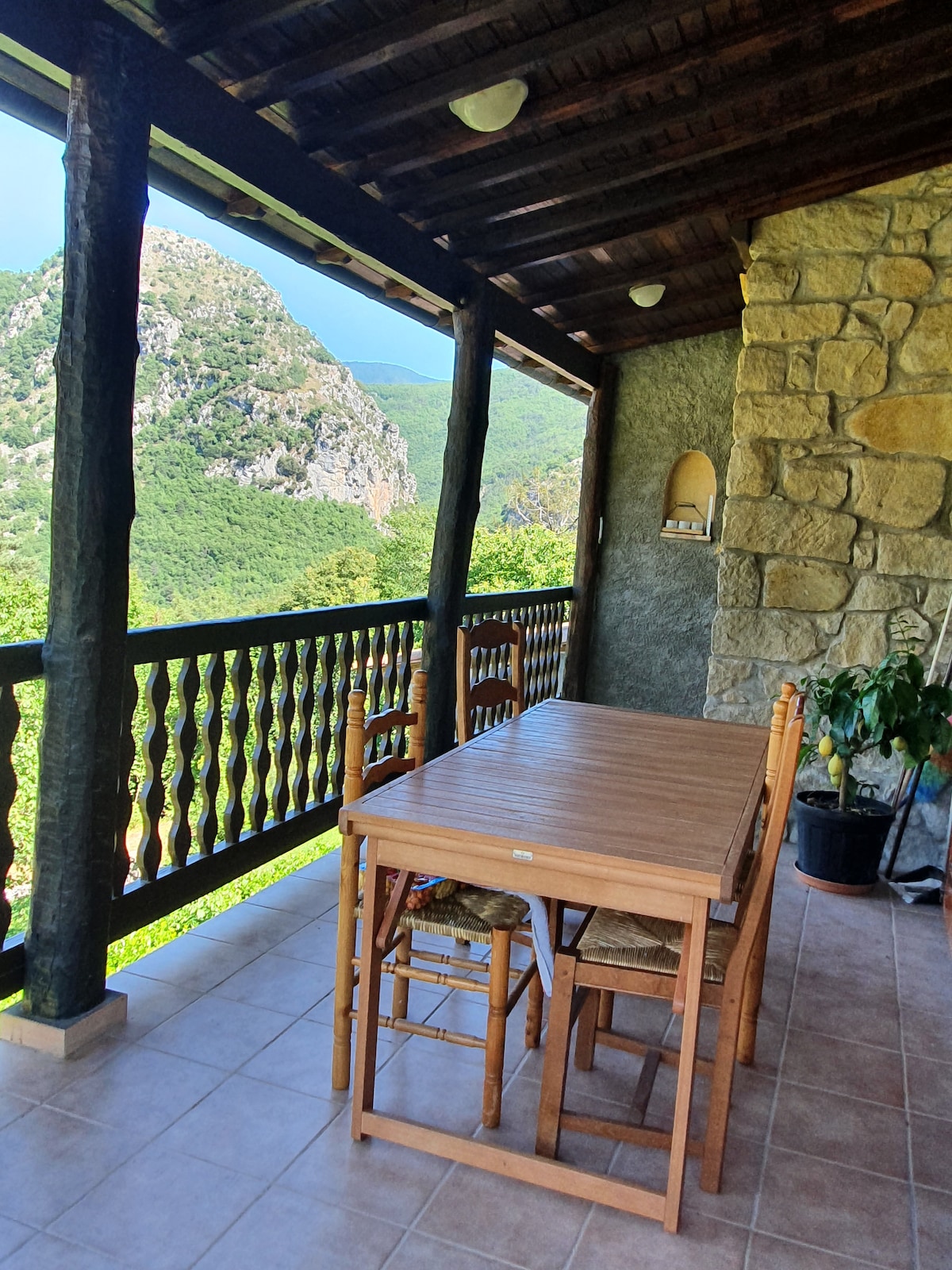 A spacious terrace features a wooden dining table surrounded by four chairs. Natural stone walls and a wooden ceiling provide a rustic ambiance. Lush green hills are visible in the background through the balcony's railings, creating a serene setting for outdoor dining.