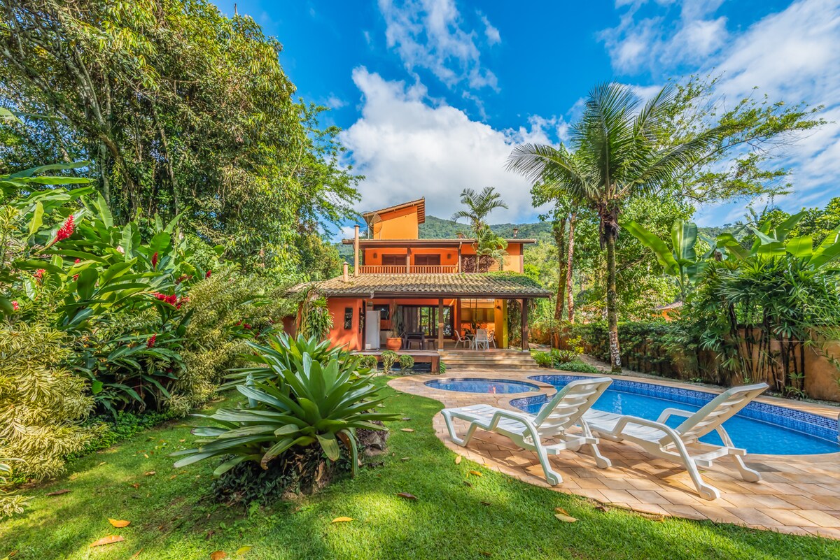 A spacious outdoor area is highlighted by a swimming pool, surrounded by well-maintained greenery and tropical plants. Sun loungers are positioned to invite relaxation. The vibrant orange house stands in the background under a clear blue sky, complemented by a mix of lush foliage.