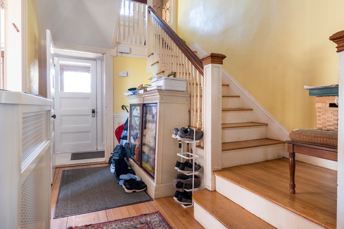 An inviting entryway is presented with yellow walls and wooden accents. A staircase leads to the upper level, while a door opens to the exterior. A shoe rack stands nearby, surrounded by various pairs of shoes. Natural light enters through the front door, enhancing the warm atmosphere.