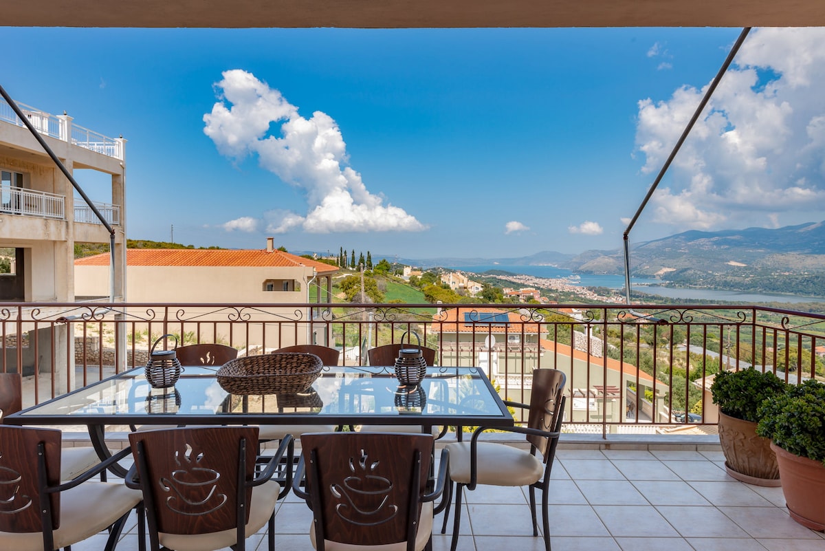 An outdoor dining area is visible on the balcony, featuring a table set with decorative elements and several chairs. The panoramic view includes distant hills and the glistening gulf, framed by a clear blue sky with a few scattered clouds.