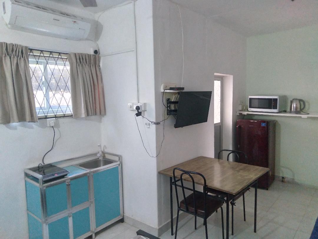 A functional kitchen area is visible, featuring a compact countertop with a sink and cooking space. A wooden dining table with black chairs is positioned nearby. A dark-colored refrigerator stands against the wall, accompanied by a microwave and kettle on a shelf. Air conditioning is also present.