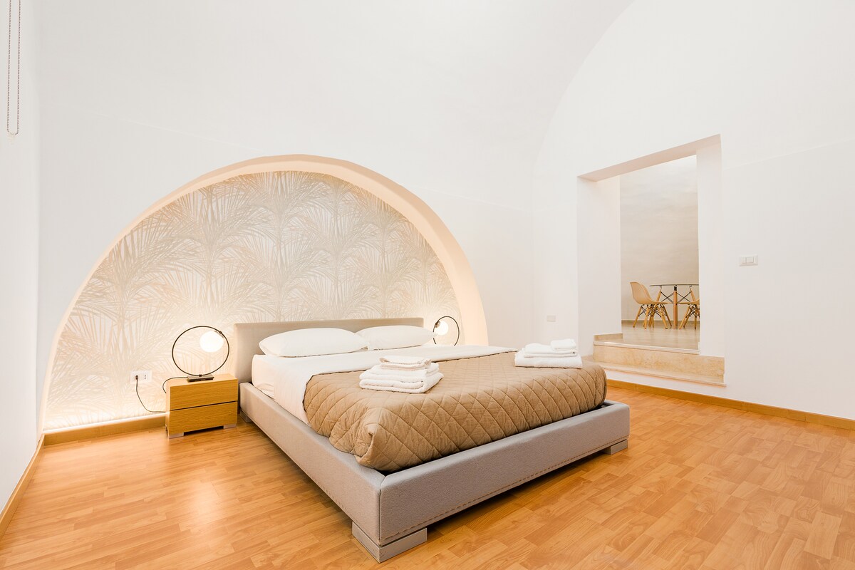 A spacious bedroom features a large bed with a neutral-toned quilt and white linens, illuminated by modern bedside lamps. An archway reveals a small seating area with a table and chairs, while warm wood flooring complements the light-coloured walls.