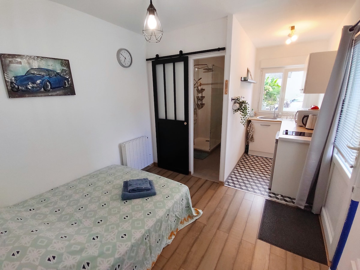 A compact studio is shown, featuring a spacious bed dressed in a patterned blanket. A modern kitchen area is visible in the background, and a sliding black door separates the bathroom. Natural light filters in through a window, illuminating the clean, simple decor.