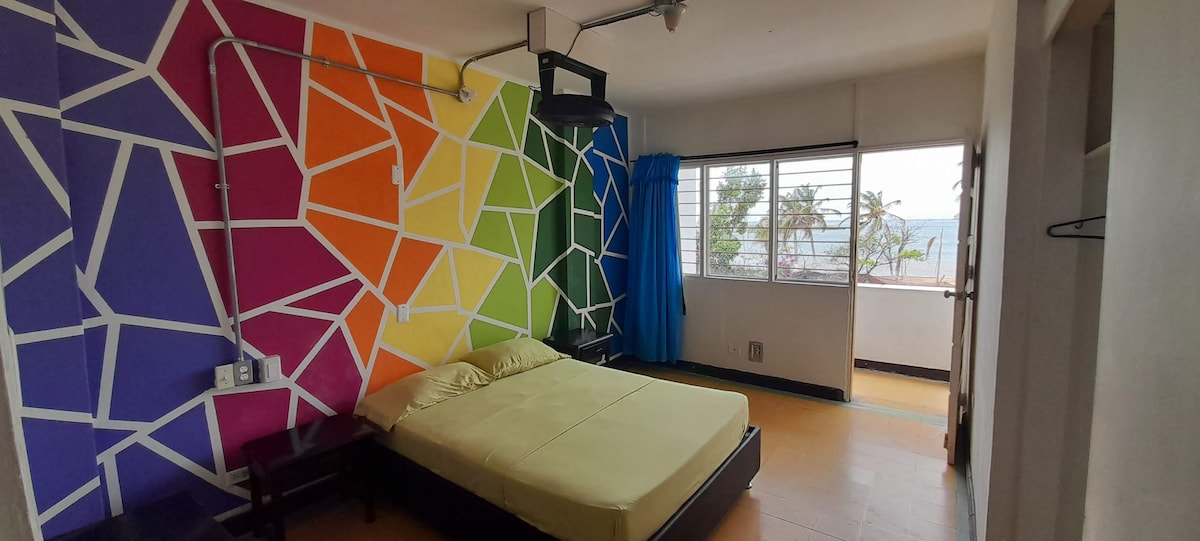 A bright room features a colorful geometric wall design in shades of pink, orange, green, and blue. A spacious bed is positioned against the wall, with natural light filtering through a large window that offers a view of palm trees and the sea. Curtains frame the window.