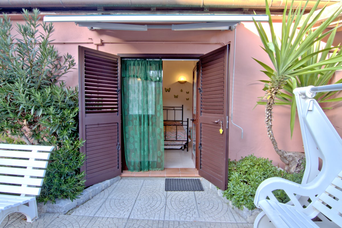 A welcoming entrance showcases open double doors with wooden shutters, leading into a tastefully decorated interior. Lush green plants frame the pathway, while a comfortable lounge chair sits nearby, inviting relaxation. Soft light is filtered through a sheer curtain, enhancing the entryway's inviting atmosphere.