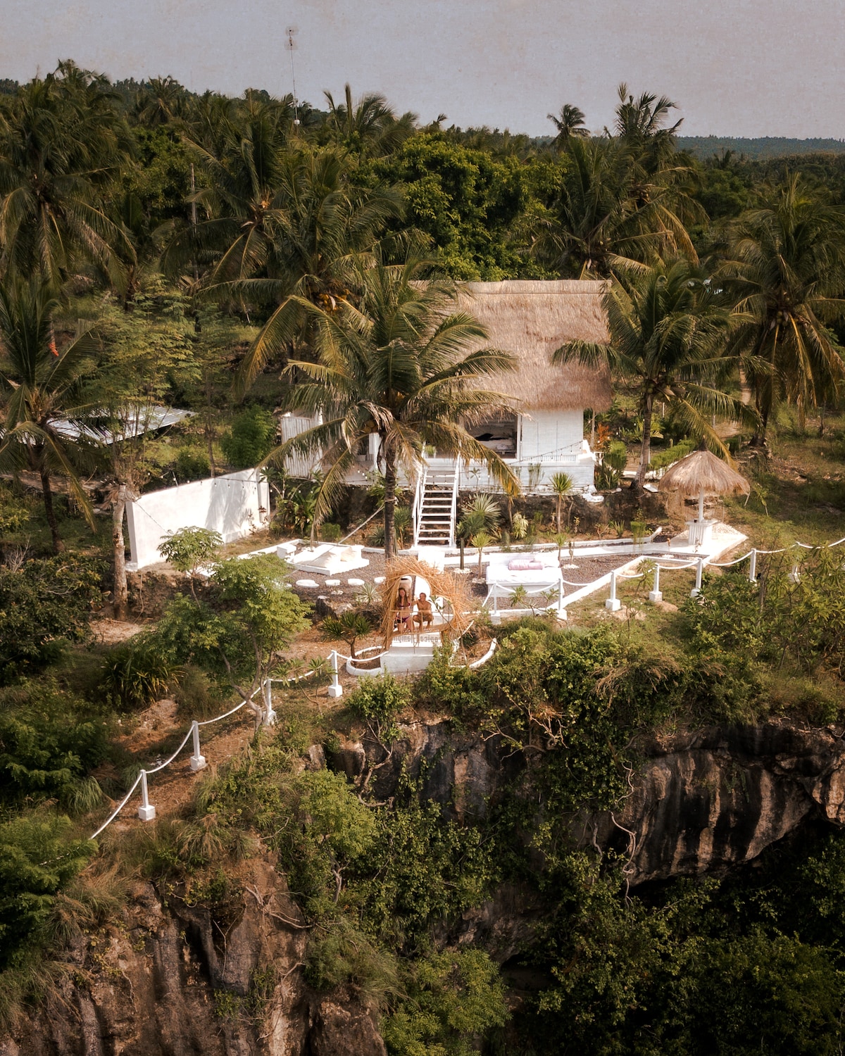 An aerial view of a secluded bamboo bungalow perched on a cliff, surrounded by lush palm trees. Outdoor lounging areas with minimalist decor are visible, along with stair access leading up to the bungalow, showcasing the vast greenery and ocean beyond.