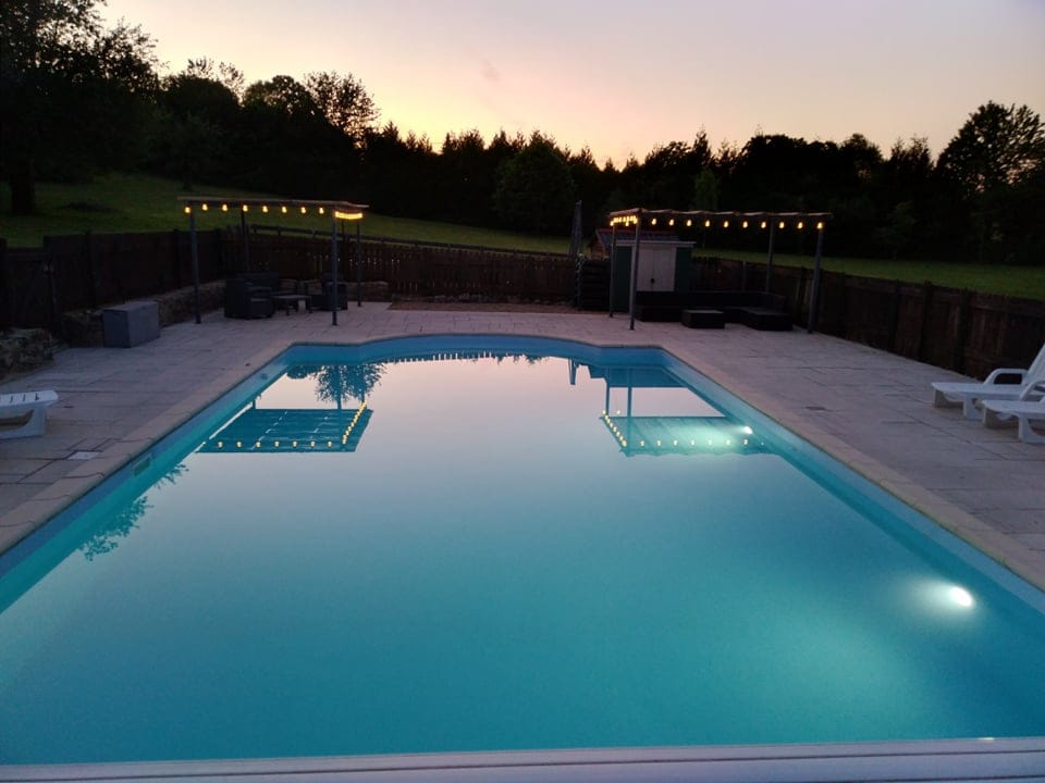A spacious outdoor pool is framed by clean, tiled surfaces and surrounded by wooden fences. Soft lights are strung above, creating a serene evening ambiance. Lush greenery is visible in the background, adding to the tranquil environment.
