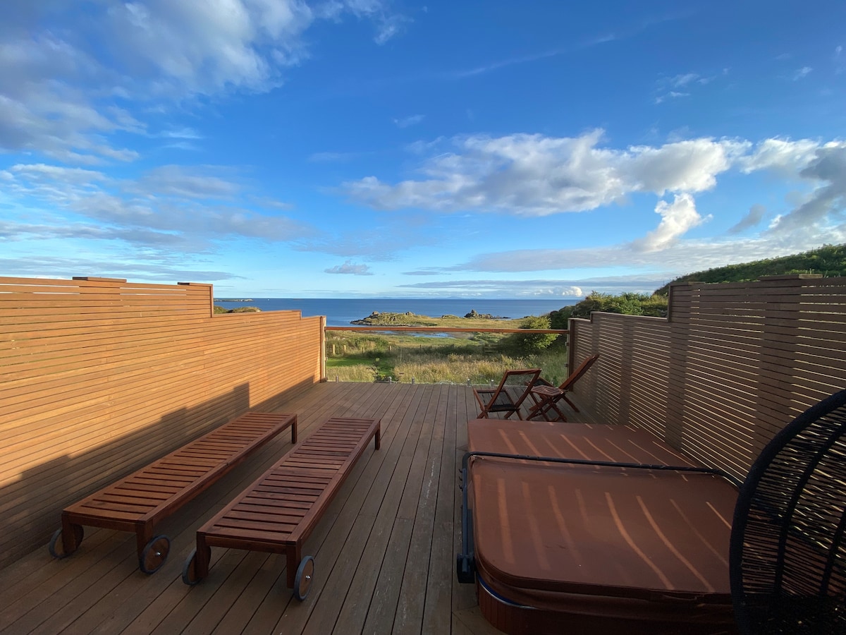 An outdoor deck features two lounge chairs and an inviting hot tub, surrounded by wooden privacy screens. The expansive ocean view stretches out in the distance, complemented by a clear blue sky filled with scattered clouds.