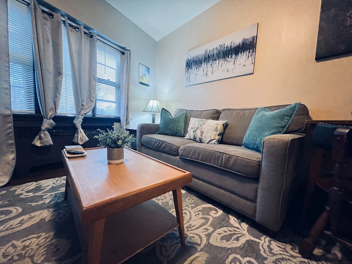 A cozy living area is presented with a comfortable gray sofa adorned with soft teal cushions. A wooden coffee table sits centrally on a decorative rug. Light filters through the window, complemented by light curtains, enhancing the room's welcoming atmosphere.