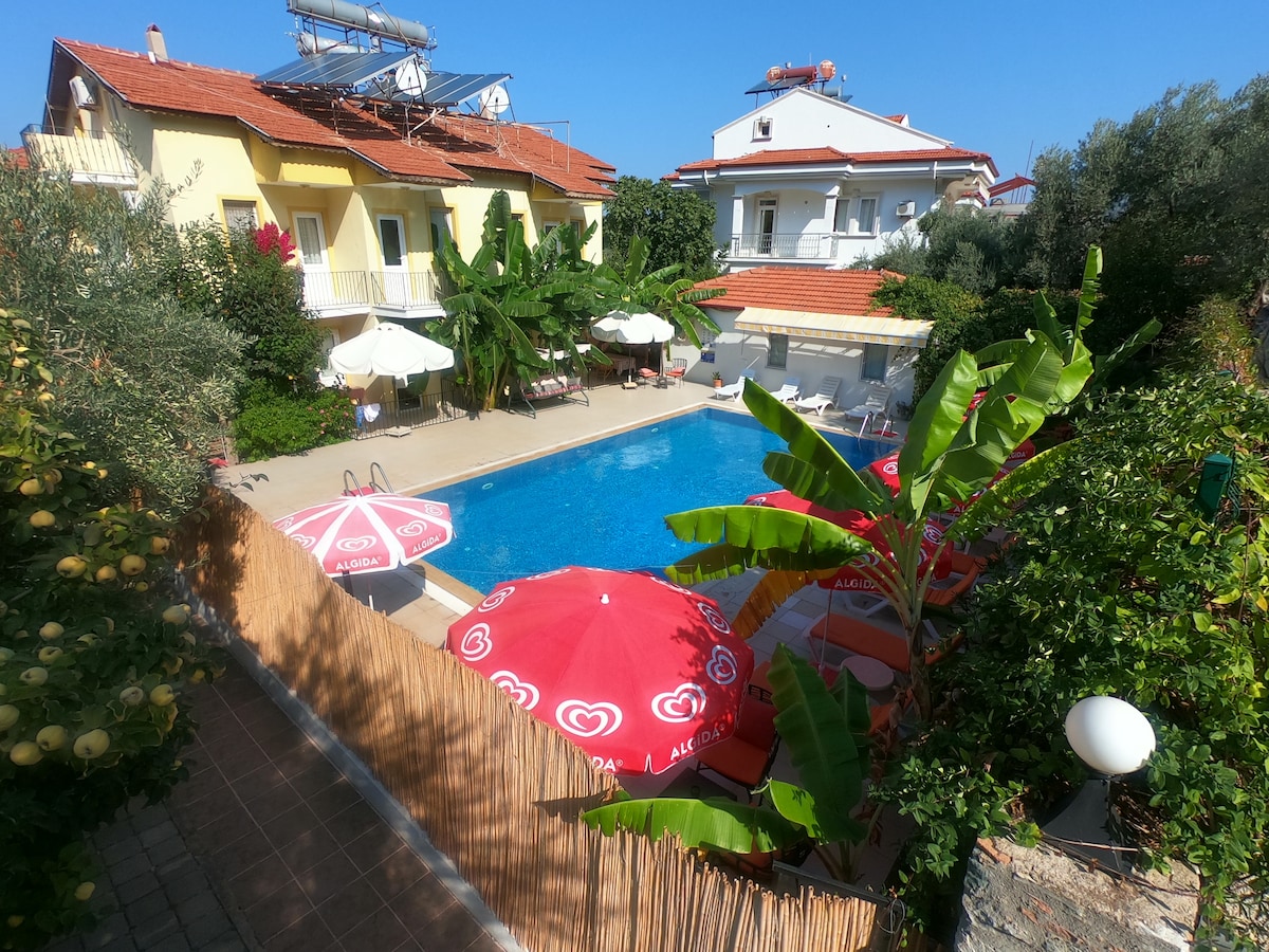 A spacious outdoor area features a private pool surrounded by umbrellas and lounge chairs. Lush greenery and palm trees create a relaxed atmosphere, while a well-kept stone path leads to the poolside. The building structure is visible in the background with a clear blue sky above.