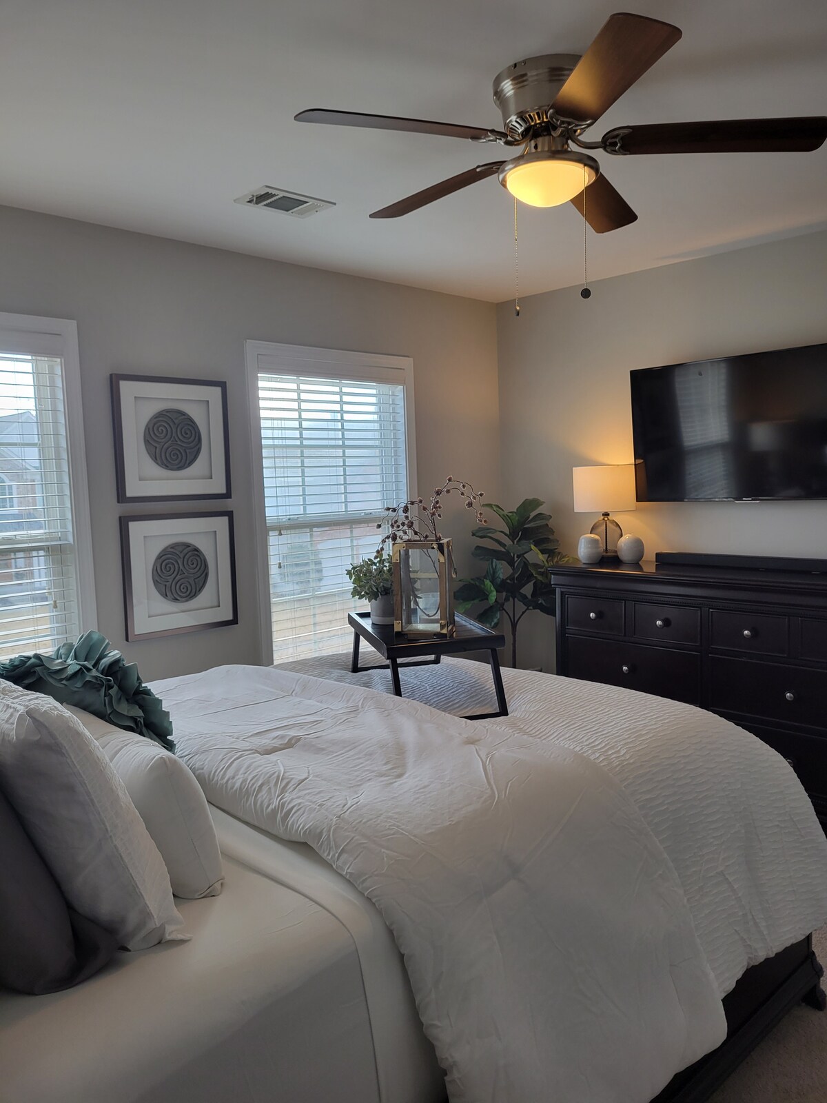A cozy bedroom features a light-colored bed dressed in white linens, accompanied by decorative pillows. A bedside table holds a lamp and decorative items. Natural light filters through two windows with blinds, enhancing the relaxed atmosphere. A wall-mounted television and artwork decorate the space.