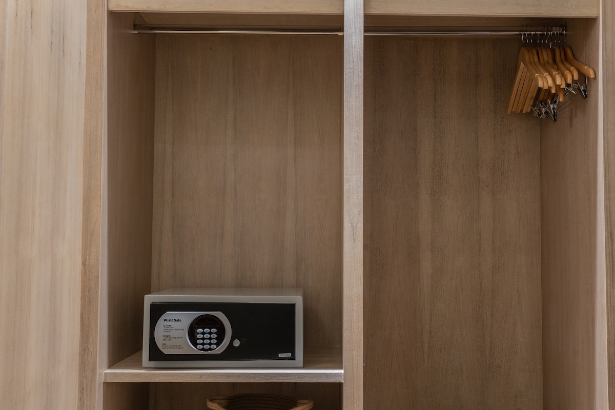 A sleek wooden wardrobe is displayed, featuring a built-in safety deposit box and several wooden hangers neatly arranged on a hanging rod. The minimalist design emphasizes functionality and security for guests' belongings.