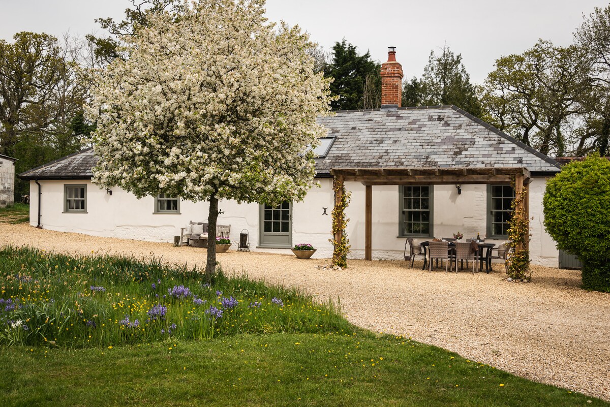 The charming exterior of the Hay Loft is showcased, featuring a whitewashed building with a slate roof. A flowering tree is positioned in front, and a gravel pathway leads to a sheltered seating area with a dining table and chairs, surrounded by green grass and blooming flowers.