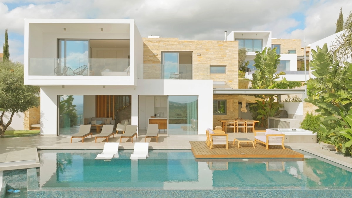 The villa's exterior showcases a modern design with large glass windows, providing views of the outdoor infinity pool flanked by sun loungers. A dining area is situated on a wooden deck, surrounded by lush greenery. The backdrop features the villa's multi-level structure and serene landscape.