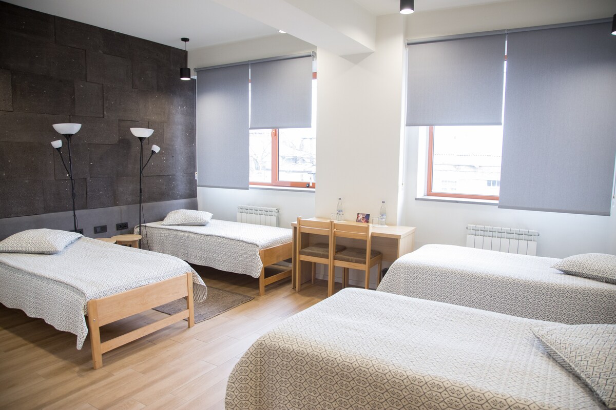 A bright guestroom features four single beds arranged in a spacious layout. Each bed is complemented by light bedding and wooden frames. Natural light filters through windows covered with shades, illuminating a simple wooden desk and lamps positioned beside the beds.