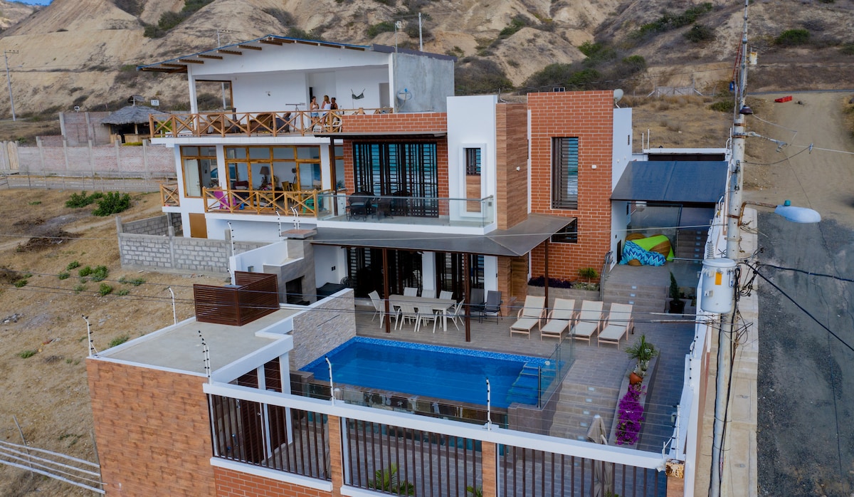 An aerial view of a new home situated near the sea, showcasing a spacious outdoor area with a rectangular swimming pool. The design features wooden balconies and large windows, complemented by a paved terrace surrounded by seating and greenery.