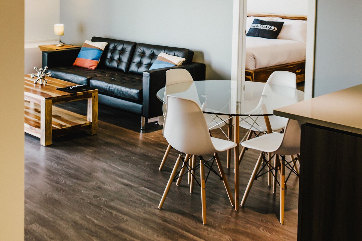 A modern living area features a sleek black couch paired with a wooden coffee table. In the foreground, a round glass dining table is surrounded by four white chairs. A glimpse of a bedroom is visible in the background, with a neatly made bed.