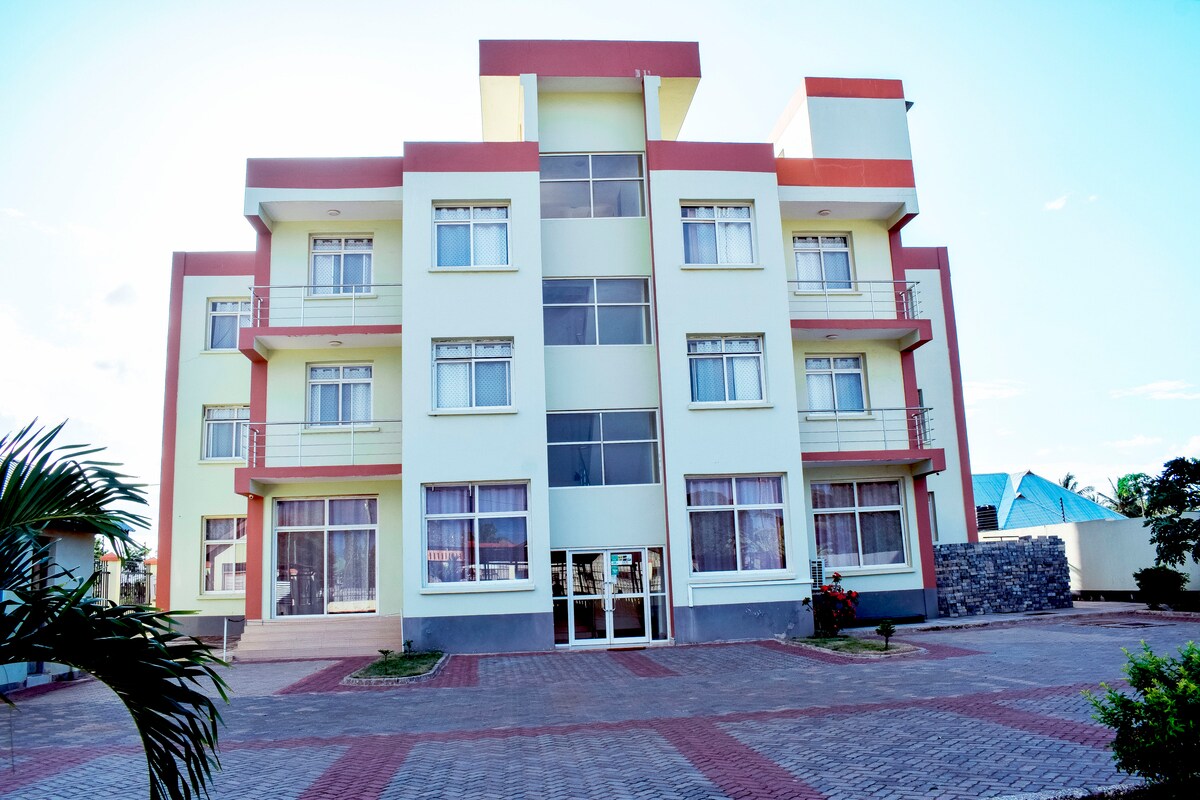 The modern three-story building features a combination of colored facades and large glass windows. A neatly paved pathway leads to the entrance, surrounded by landscaping with small plants. The structure presents a clean and contemporary look in an upscale environment.