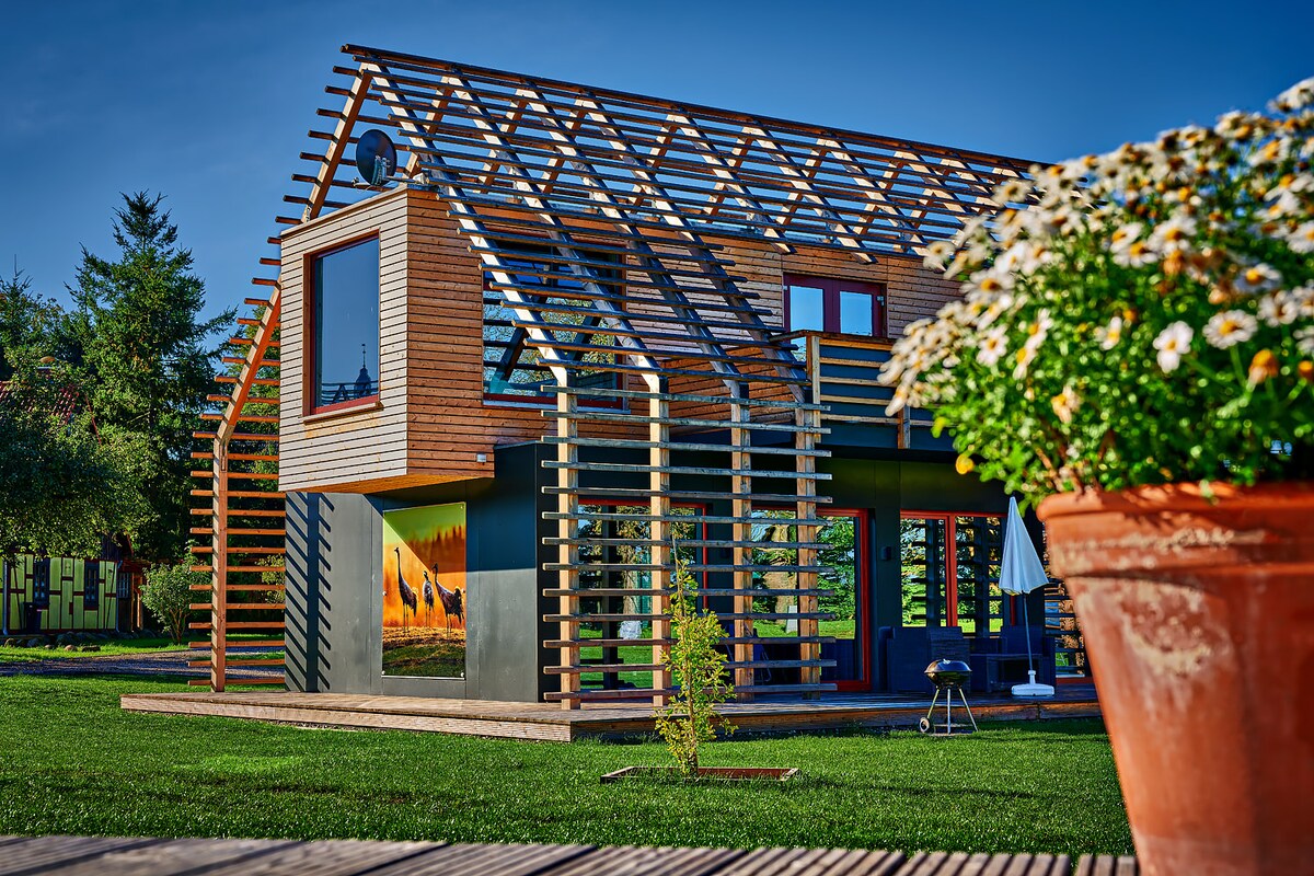 A contemporary chalet structure showcases a unique wooden frame design, allowing for ample natural light. The exterior features large windows and a porch area surrounded by well-maintained greenery. A potted flower arrangement is positioned in the foreground, adding a touch of nature.