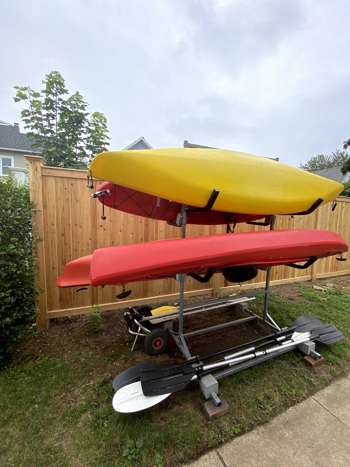 A kayak rack holds three kayaks in vibrant colors: yellow at the top, followed by red in the middle, and another red kayak at the bottom. Paddles are positioned beneath the kayaks, and a wooden fence lines the background, creating a private outdoor space.