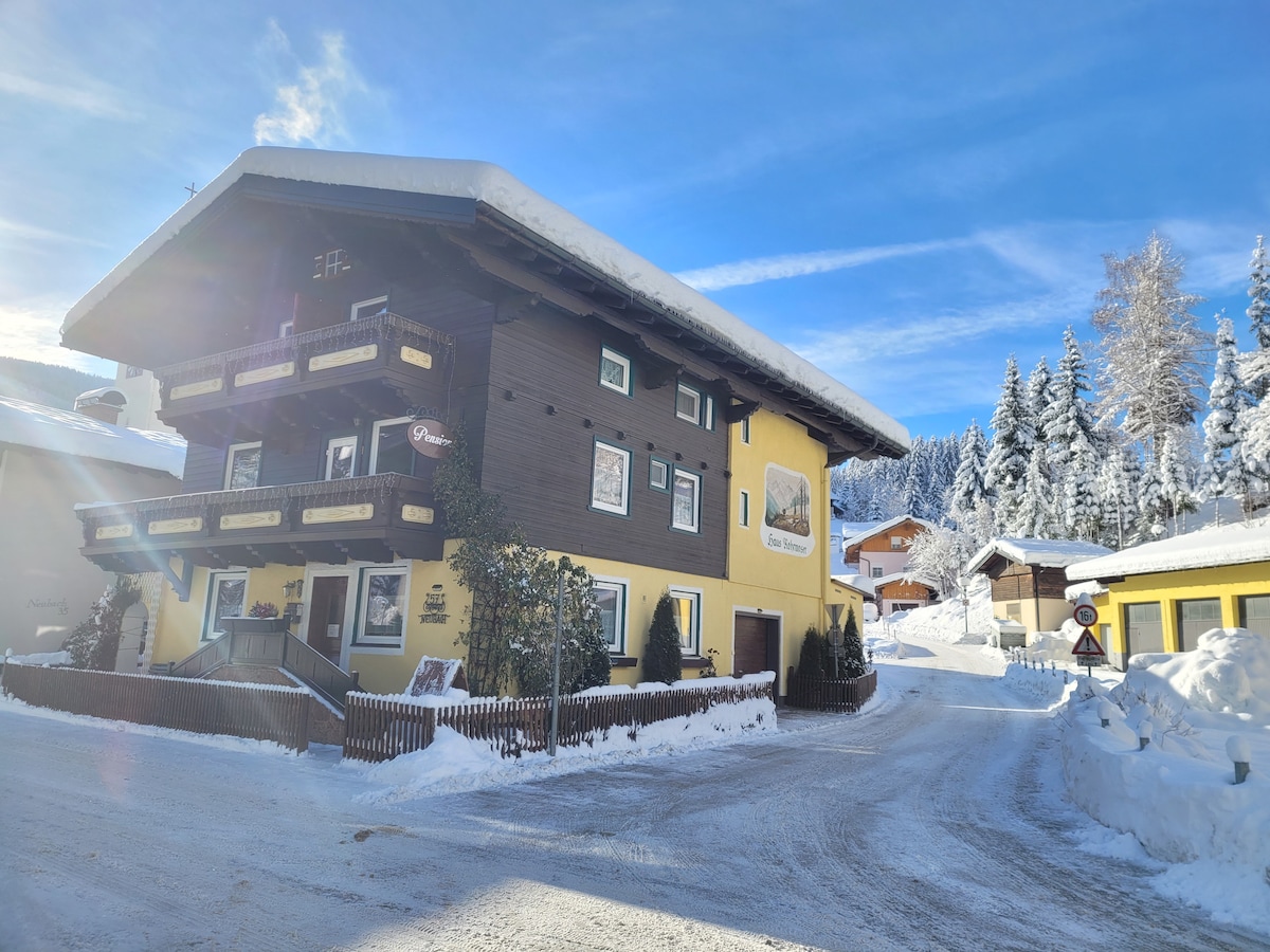 The charming guesthouse is framed by snow-covered trees, showcasing a two-story structure with wooden balconies and a bright yellow facade. A sloped, snowy driveway leads to the entrance, offering a welcoming atmosphere in the winter landscape.