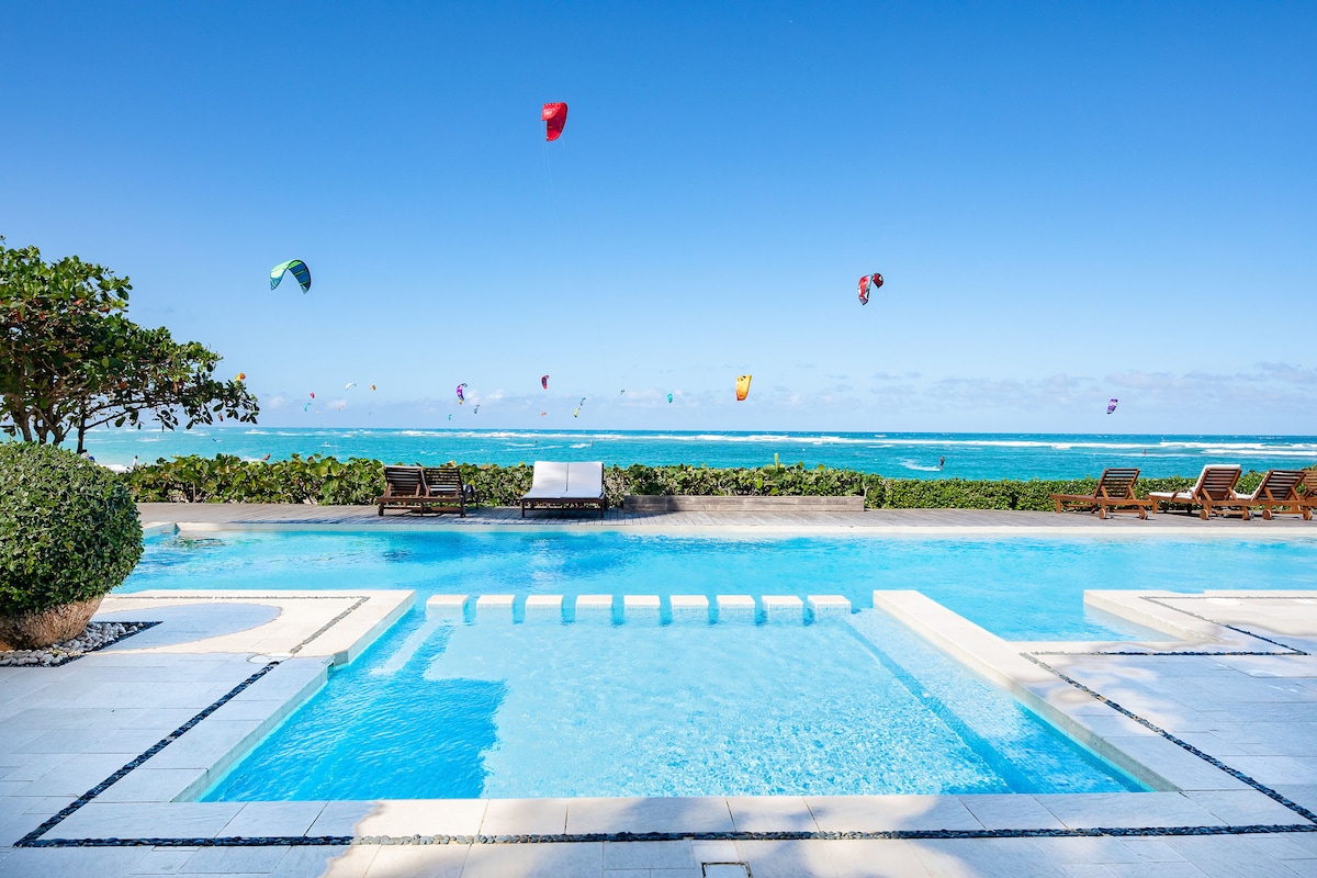 A large, clear pool is situated in the foreground, bordered by stone tiles and surrounded by lush greenery. Kitesurfers can be seen gliding over the vibrant blue ocean in the background, under a bright sky with scattered clouds.
